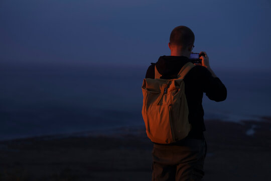 back rear view of one alone backpacker Man with rucksack, cell phone standing on peck of rock, taking photos of the view. Man on top of a mountain photographing sky above Black sea or pacific ocean.