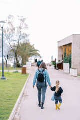 Mother and her little daughter are walking along the road in a green park. Back view