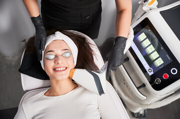 Top view of cheerful woman in safety goggles receiving laser facial treatment in cosmetology...