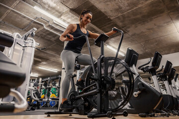 A sportswoman is doing exercises on an air bike in a gym.