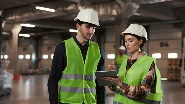 Two industrial engineers in safety gear engaged in a professional discussion over a digital tablet in a warehouse setting, exemplifying teamwork and technology in industry