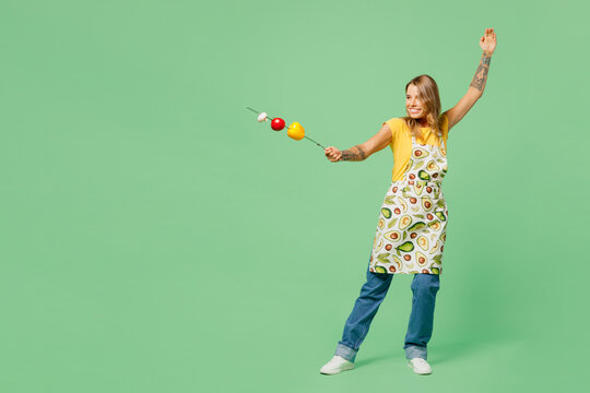 Full body fun young housewife housekeeper chef cook baker woman wear apron yellow t-shirt hold skewer with grilled vegetables pov fight isolated on plain green background. Cooking vegan food concept.