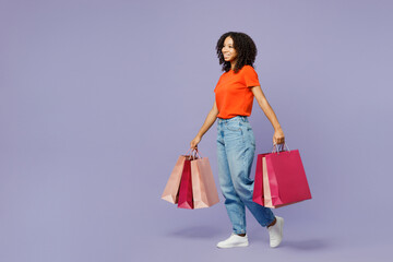 Full body little kid teen girl of African American ethnicity wear orange t-shirt hold paper package bags after shopping isolated on plain purple background studio. Black Friday sale buy day concept.