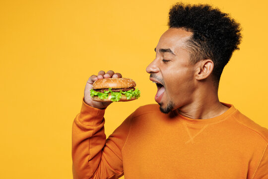 Close up young man of African American ethnicity wear orange sweatshirt casual clothes hold eat bite fast food burger open mouth isolated on plain yellow background studio portrait. Lifestyle concept.