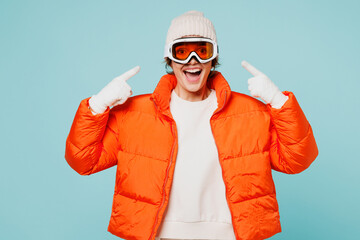 Young smiling fun woman wear warm padded windbreaker jacket hat ski point finger on goggles mask looking camera travel rest spend weekend winter season in mountains isolated on plain blue background © ViDi Studio