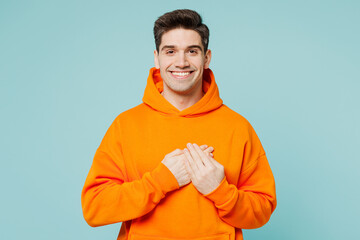 Young smiling happy cheerful man he wear orange hoody casual clothes put folded hands on heart look...