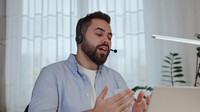 Focused young businessman in headset holding video call on laptop and answering client's questions in home interior. Concentrated male consulting customer online and presenting product for sale.