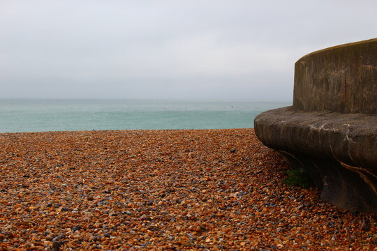 Empty pebble beach on England's south coast on an overcast, rainy, grey, summer day with cloudy skies, a rough see, and an empty cement curved bench. Beach with rocks and waves and ample copy space