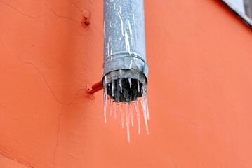 Downspout with icicles on red wall background. Icicles hanging from gutter. Ice melt in drain...