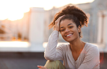 Young happy african american girl looking at camera while sitting on roof terrace on sunny morning