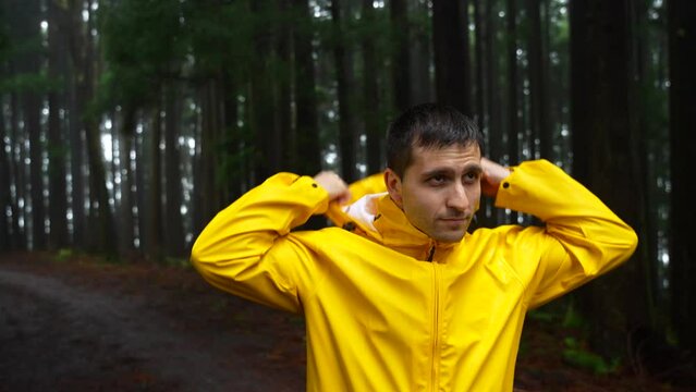 Young man zipping bright yellow raincoat in dark rainy forest, looking around