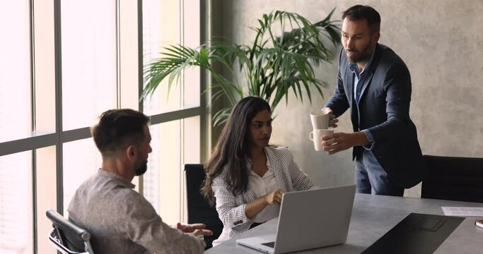 Middle-aged colleague in suit bringing tea cups, morning beverage to multiethnic teammates, team members having coffee break during workday gathered in modern conference room. Pause, friendly relation