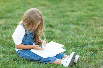 Cute little girl sitting on the grass and reading a book on summer day. Children education concept.