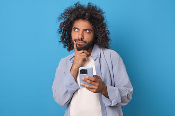 Young thoughtful attractive Arabian man with phone in hands comes up with new post for page on social network and touches face and looks to side choosing words for message stands on blue background.