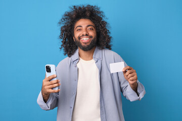 Young cheerful handsome Arabian man with credit card and smartphone in hands laughs after making purchase and rejoices at opportunity to use online banking to make payments stands on blue background.