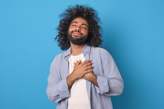Young attractive happy Arabian man puts hands on heart and with eyes closed raises head showing gratitude or empathy and wishing well to people around posing on isolated blue background