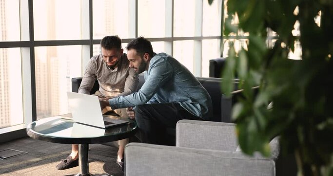 Two smiling friendly workmates working online using laptop, learn new software or corporate program sit at desk staring at laptop screen looking interested engaged in teamwork working on joint project