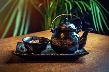 A traditional Asian ceramic teapot and a bowl with cane sugar on the table, against a background of...