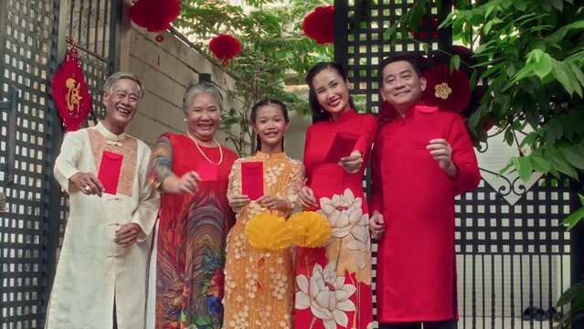 Medium long portrait of happy Asian family showing traditional red envelopes as gifts for Tet or Lunar New Year to camera outdoorsMedium long portrait of happy Asian family showing traditional red env