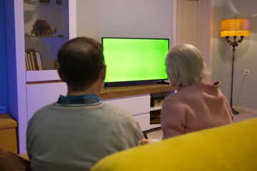 Family watching TV. Green screen. An adult man and his elderly mother are sitting at home on the couch and watching TV. In front of them is a green screen TV. 
 - obrazy, fototapety, plakaty
