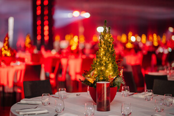  table with a lit Christmas tree centerpiece in a banquet hall with red ambient lighting and blurred background tables.