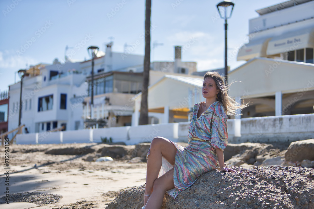 Wall mural Young and beautiful woman in a sequined dress and high heels, sitting on a large rock on the beach, happy, relaxed and calm. Concept beauty, fashion, trend, peace, relaxation.