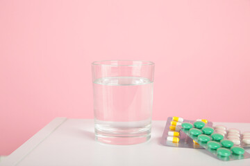 Pink alarm clock with glass of water and blister packages of pills on light table background.