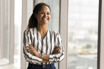 Happy beautiful young African professional woman in casual shirt posing in office with arms...