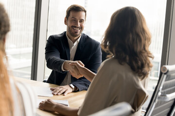 Positive millennial business partners shaking hands on corporate meeting. Confident businessman giving greeting, handshake to female colleague over table, smiling, laughing