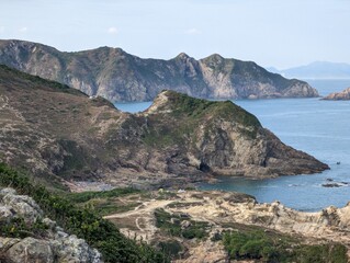 A scenic view of a coastline with mountains and a sea. The foreground has trails and grass, the middle ground has rocky hills, and the background has lush mountains.