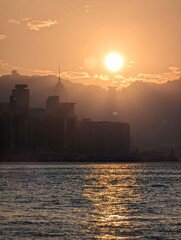 Fototapeta premium A serene sunset over a cityscape and water. The sun casts a golden hue and reflects on the water. The city silhouette shows skyscrapers and mountains.