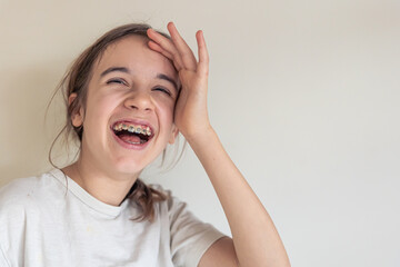 A teenage girl with braces smiles against the background of a white wall.