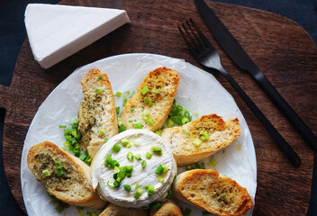 Brie cheese with green onions with bread croutons on a plate next to cutlery on a dark background