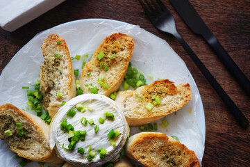 Brie cheese with green onions with bread croutons on a plate next to cutlery on a dark background