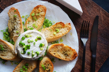 Brie cheese with green onions with bread croutons on a plate next to cutlery on a dark background