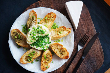 Brie cheese with green onions with bread croutons on a plate next to cutlery on a dark background
