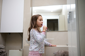 Little kid girl brushing her teeth, standing in the gray minimalist home bathroom. Dental care. Oral hygiene. Copy space