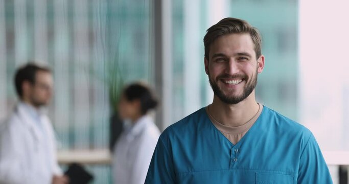 Serious young handsome doctor man in blue uniform shirt getting cheerful, happy, looking at camera with toothy smile, standing in hospital hall space with colleagues in background