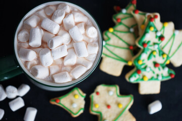 Cocoa with marshmallows next to cookies in the shape of Christmas trees on a black background