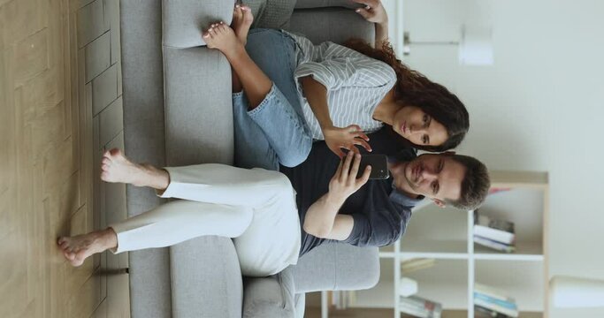 Serious young couple using ecommerce application, Internet technology on smartphone together, sitting with cellphone in sofa, shopping online, talking. Vertical full length shot