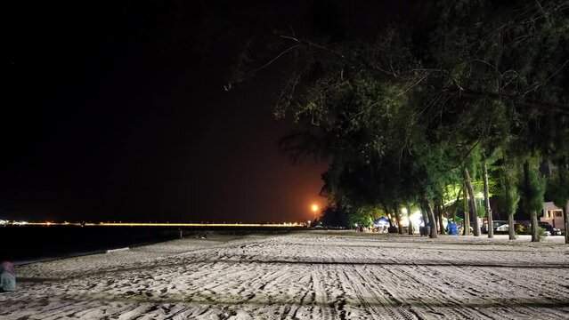 Nighttime at Pantai Puteri Beach in Melaka. A refinery tower at the horizon emits fluctuating lights. Traffic flows on the road behind the beach's illuminated trees lit by street lamps.