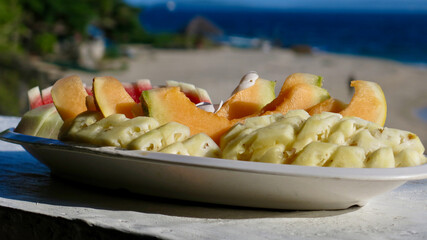 Fruit plate on the beach. Sliced fruits on a white plate against the background of the sea and the beach.