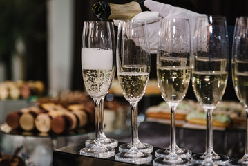 Catering service concept. The waiter pouring white sparkling wine. Barmen pours champagne into flute glasses. Champaign is being poured into glasses. Bottle in a closeup view. Rows of full glasses. - obrazy, fototapety, plakaty