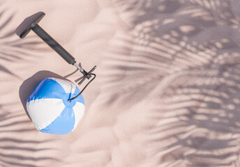 blue and white beach ball connected to a black pump on sandy texture with palm tree shadows, beach leisure concept.