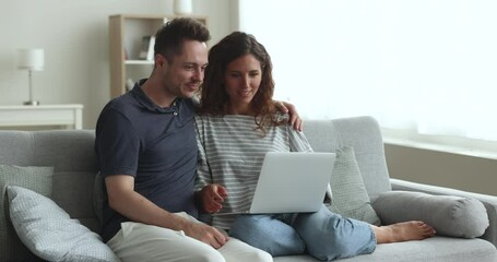 Positive young romantic couple holding computer, relaxing on comfortable couch at home, watching video, movie on laptop together, using e-commerce application for shopping, travel booking - Powered by Adobe