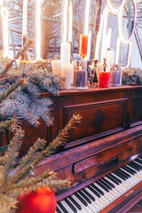 A lacquered piano made of mahogany stands in front of a Christmas tree, decorated with Christmas tree branches and various festive decorations, candles and bright warm yellow lamps, creating a festive