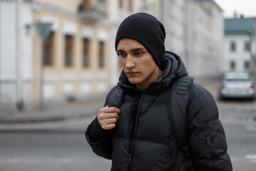 man in black winter clothes with a knitted hat, down jacket and backpack walks in the city