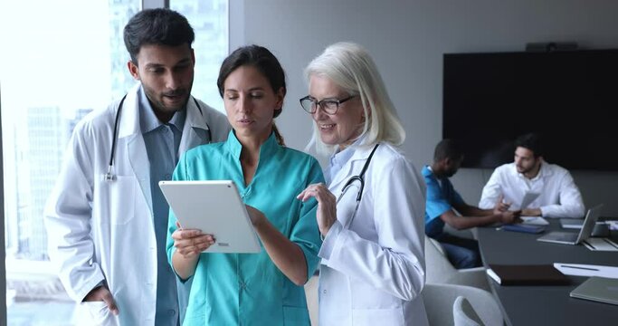 Happy diverse team of younger and elder doctors in uniforms using tablet together, browsing Internet, touching screen, looking at screen, talking, smiling, laughing. Clinic staff meeting in background