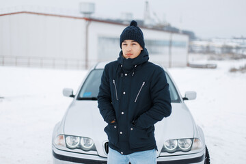 handsome man with a winter knitted hat in a stylish jacket stands near a car on a snowy winter day....