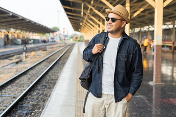 Young asian man traveler backpack in the train station. Backpacker male wear hat and denim jacket at the railway. Travel concept. The concept of a man traveling alone.